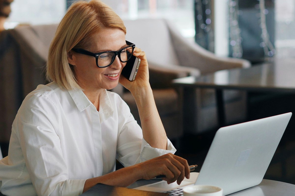 Donna di mezza età con occhiali che sorride al telefono mentre lavora al portatile in un caffè — La vera prova inizia dopo il pagamento
