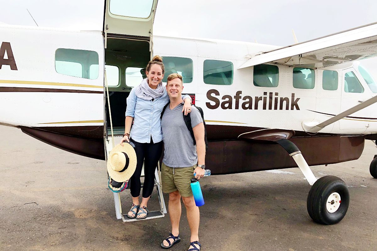 Couple souriant sur les marches d'embarquement d'un avion à hélices Safarilink, la femme tient un chapeau de paille et l'homme une bouteille d'eau — Choisissez votre style de voyage pour un safari