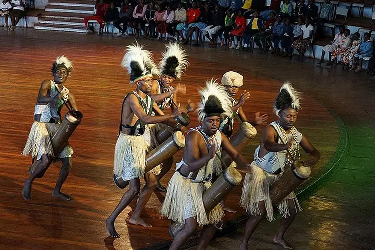 Traditionella afrikanska trummisar och dansare i fjäderprydda huvudbonader och gräskjolar som uppträder på en träarena inför sittande publik.