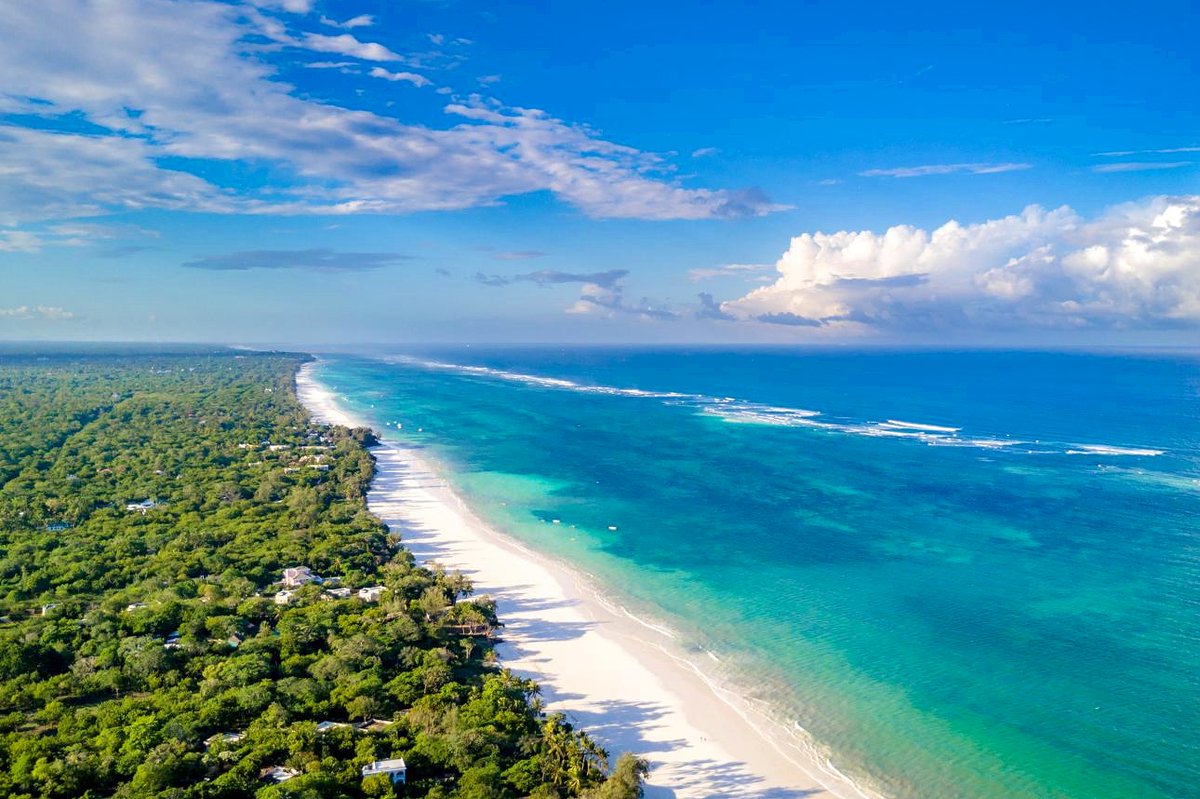 Flygfoto av en lång vit sandstrand och turkost hav intill frodig grön kustskog under en klarblå himmel — Niroskos 'Varför?' blogg.