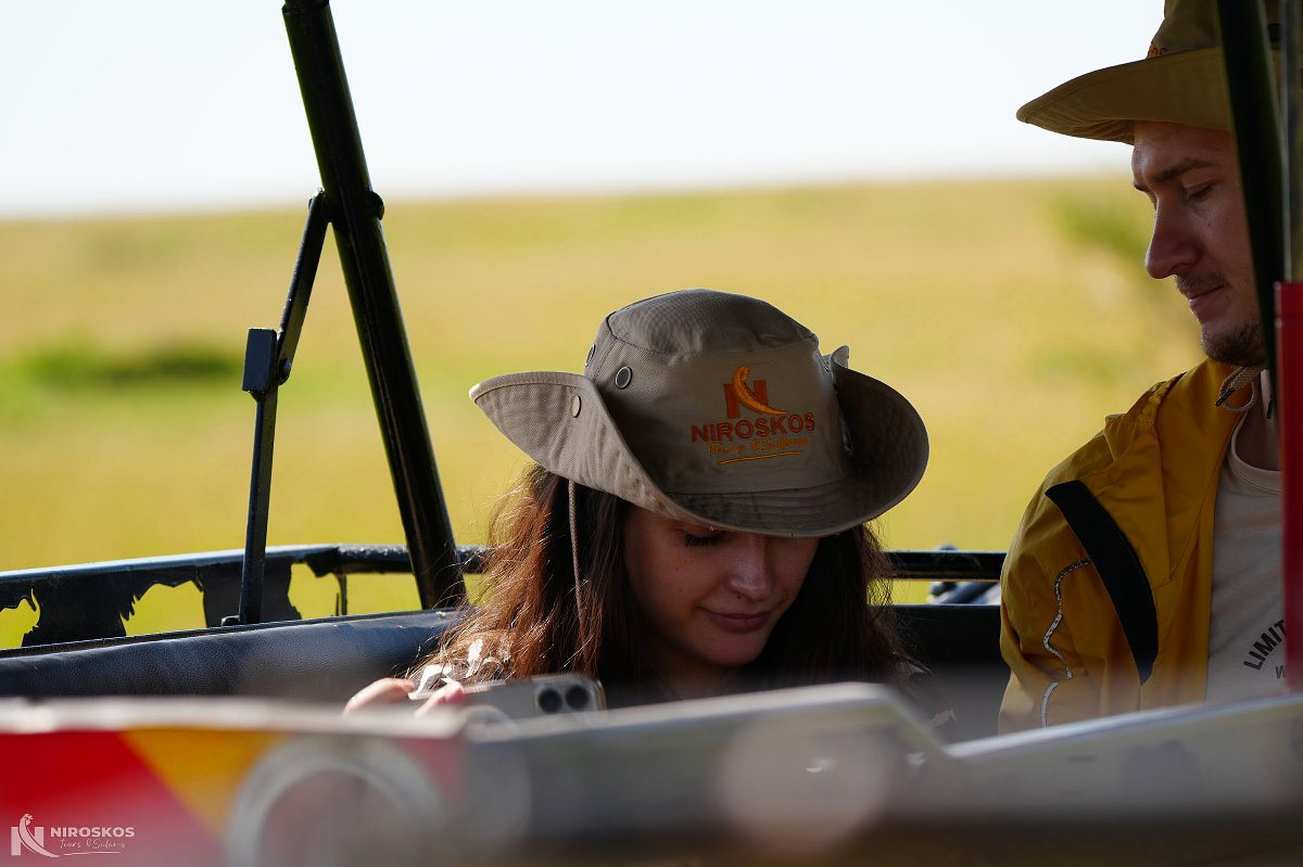 Turista con sombrero de safari de la marca Niroskos junto a un guía con chaqueta amarilla dentro de un vehículo de safari abierto, sabana dorada al fondo.
