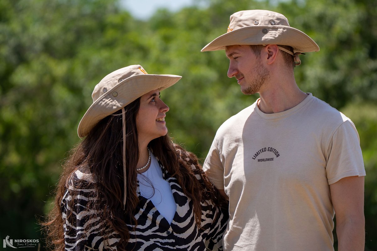 Pareja sonriente con sombreros de safari color caqui mirándose ante un fondo verde bañado por el sol — Más que un viaje