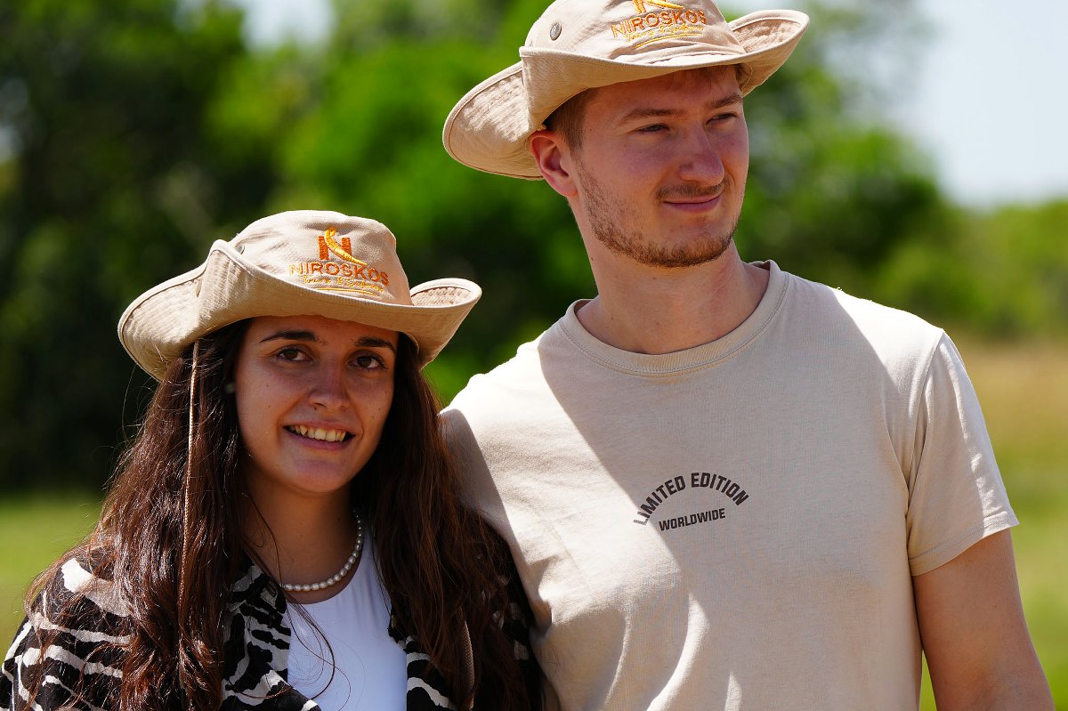 Two smiling guests wearing Niroskos safari hats at a luxury lodge, sunlit green savanna in the background
