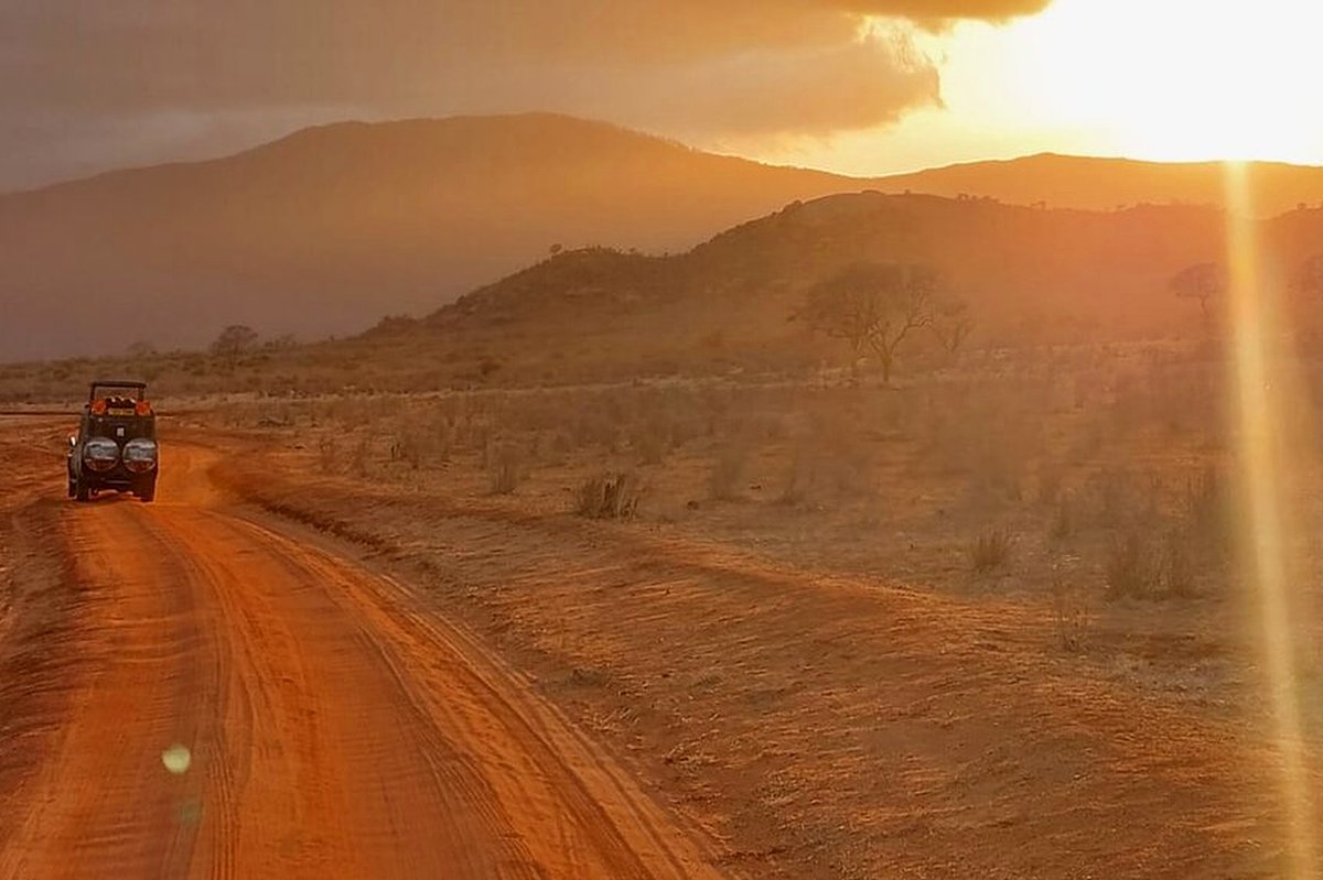 Open safari vehicle driving a red dirt track toward a glowing sunset across the Tsavo plains